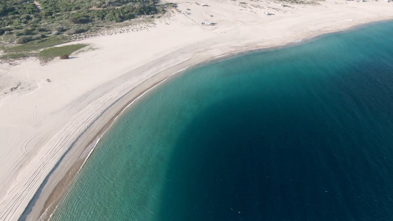 A second aerial view of Los Frailes coastline shows more of the beach arc, shallow turquoise water near the sand, darker water offshore, sparse vegetation, and no visible people.
