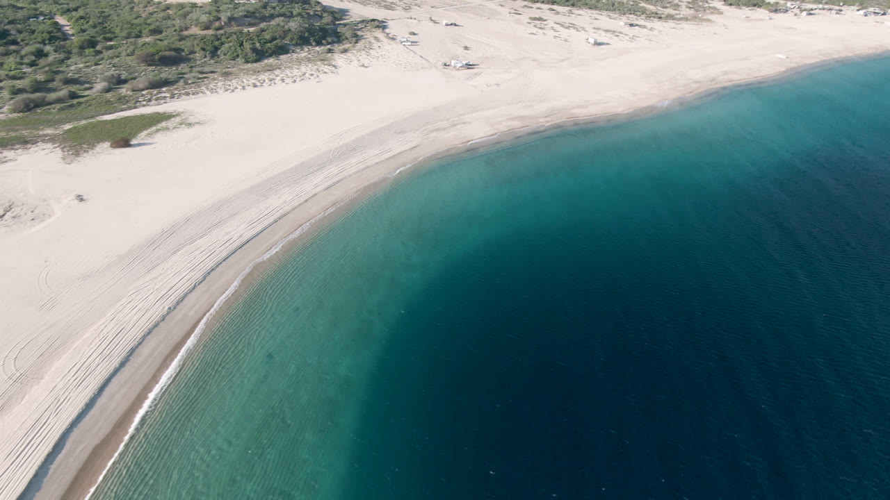 Aerial coastline frame with broad sand, low vegetation, and deep blue water.
