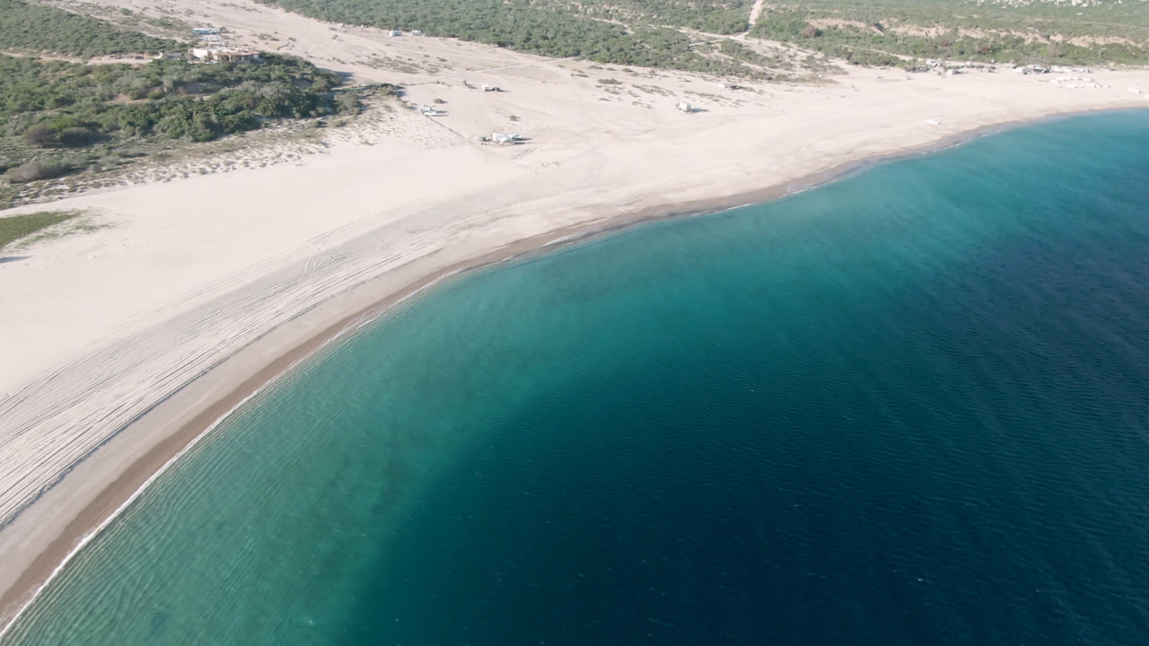 Aerial frame over Los Frailes, Baja California Sur, showing a pale curved beach, blue-green coastal water, scrub-covered land, distant hills, and no visible people.