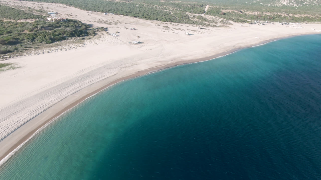 Aerial view of Los Frailes shoreline with turquoise shallows spreading into darker water, a wide pale beach, scrubland behind it, distant mountains, and no visible people.