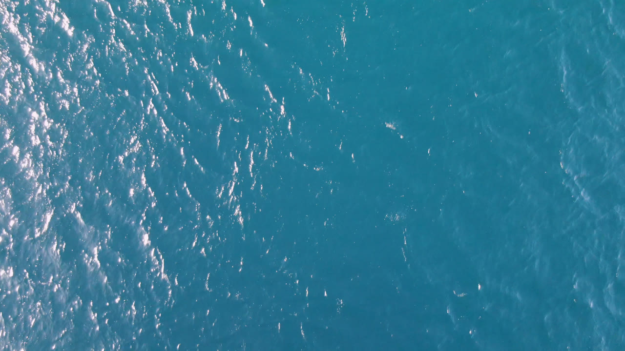 Aerial frame over blue-green water in Los Frailes, with a brighter strip of reflected sunlight along the left side and no visible people.