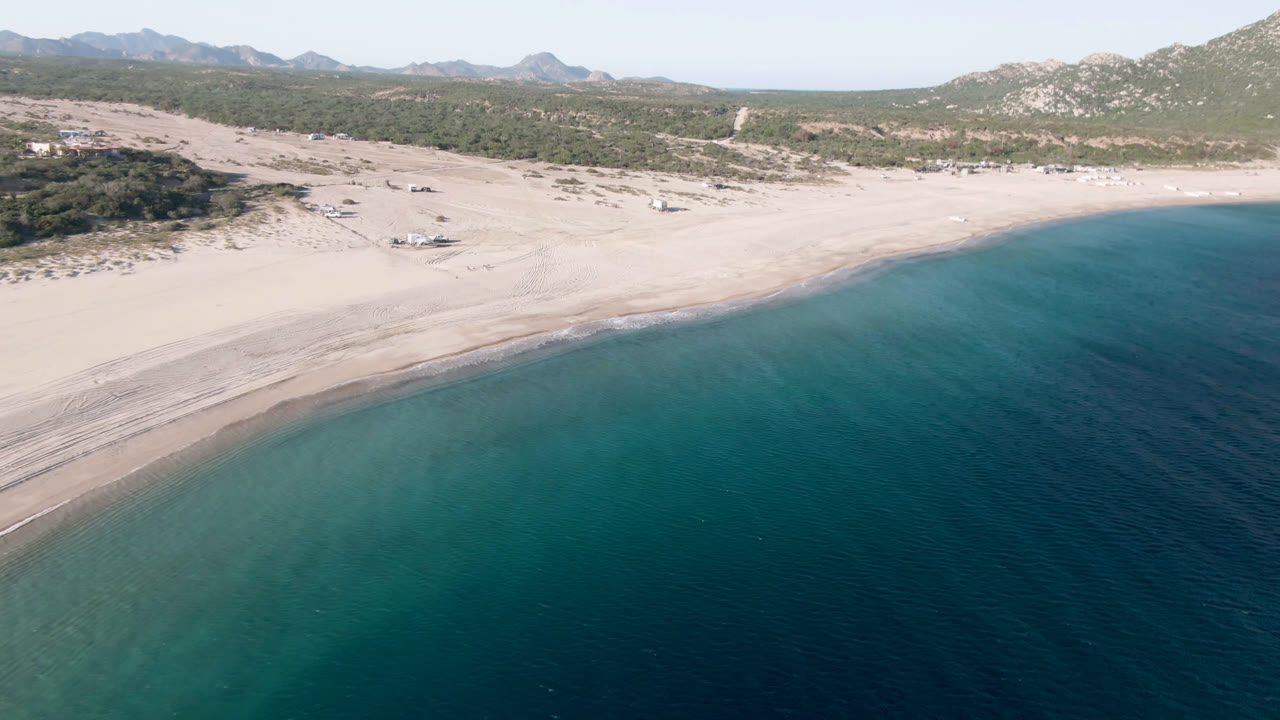 At 0:00, an aerial view over Los Frailes, Baja California Sur, Mexico shows blue-green water in the foreground, a pale sandy beach, low scrub hills, distant mountains, and no visible people.