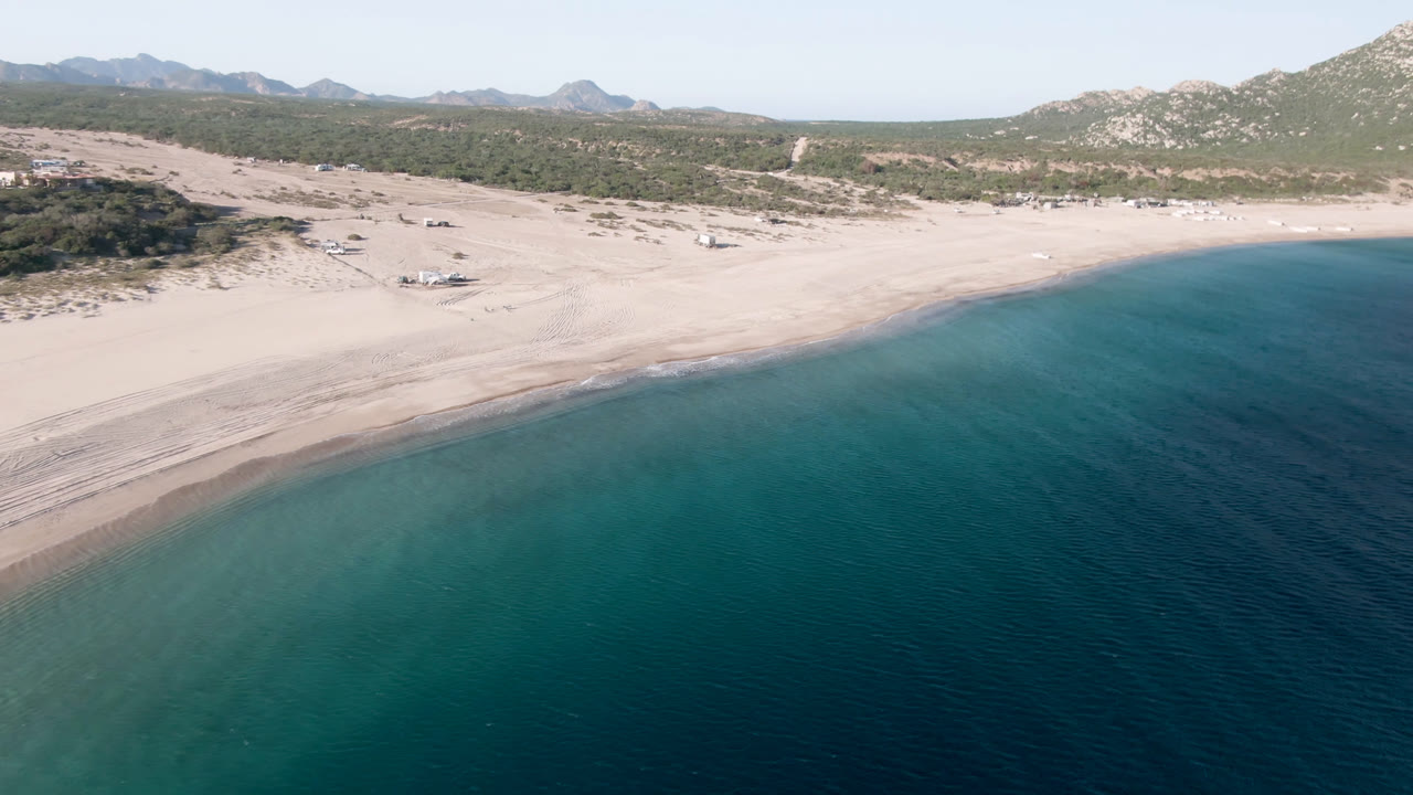 At 0:01, the drone view over Los Frailes shows the beach curve slightly farther along, with turquoise shallows, tire-like sand tracks, small boats onshore, hills behind, and no visible people.