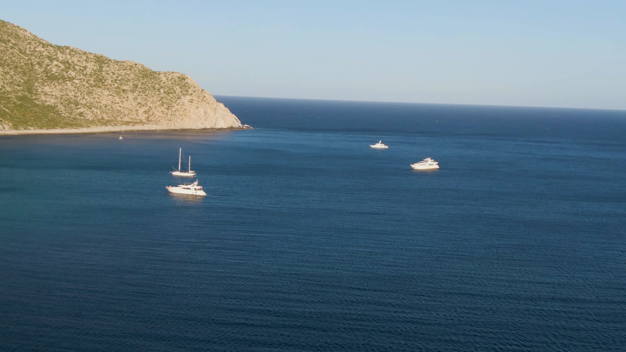 At 0:00, several white boats float on blue water off Los Frailes, Baja California Sur, with a rocky green-tan shoreline in the background and no visible people.