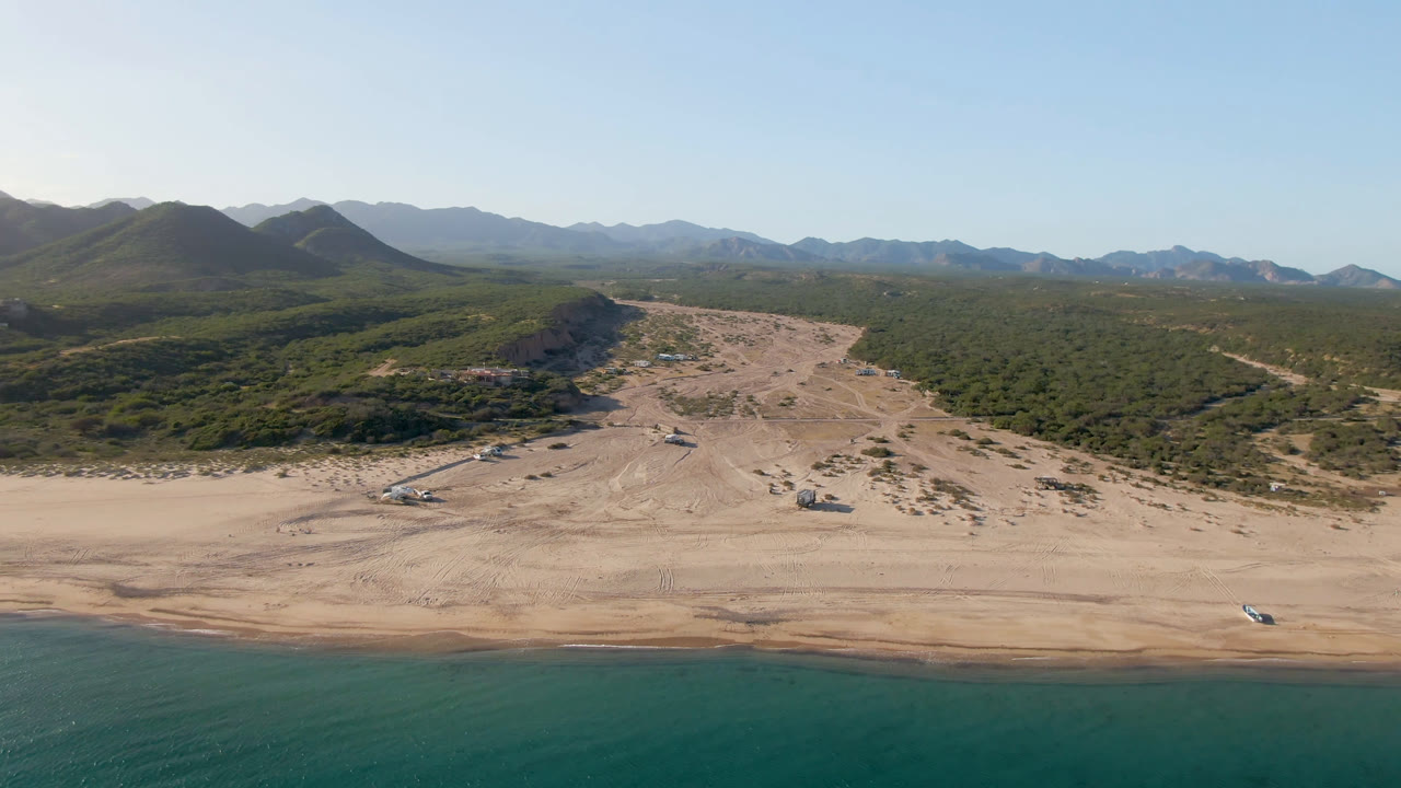 Wide aerial frame over Los Frailes with turquoise water at the lower edge, a broad sandy beach, green scrubland, distant mountains, clear sky, and no visible people.