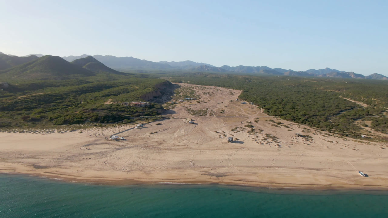 Los Frailes aerial still with the shoreline running across the lower frame, pale sand and vehicle tracks through the center, green desert vegetation behind it, mountains on the horizon, and no visible people.