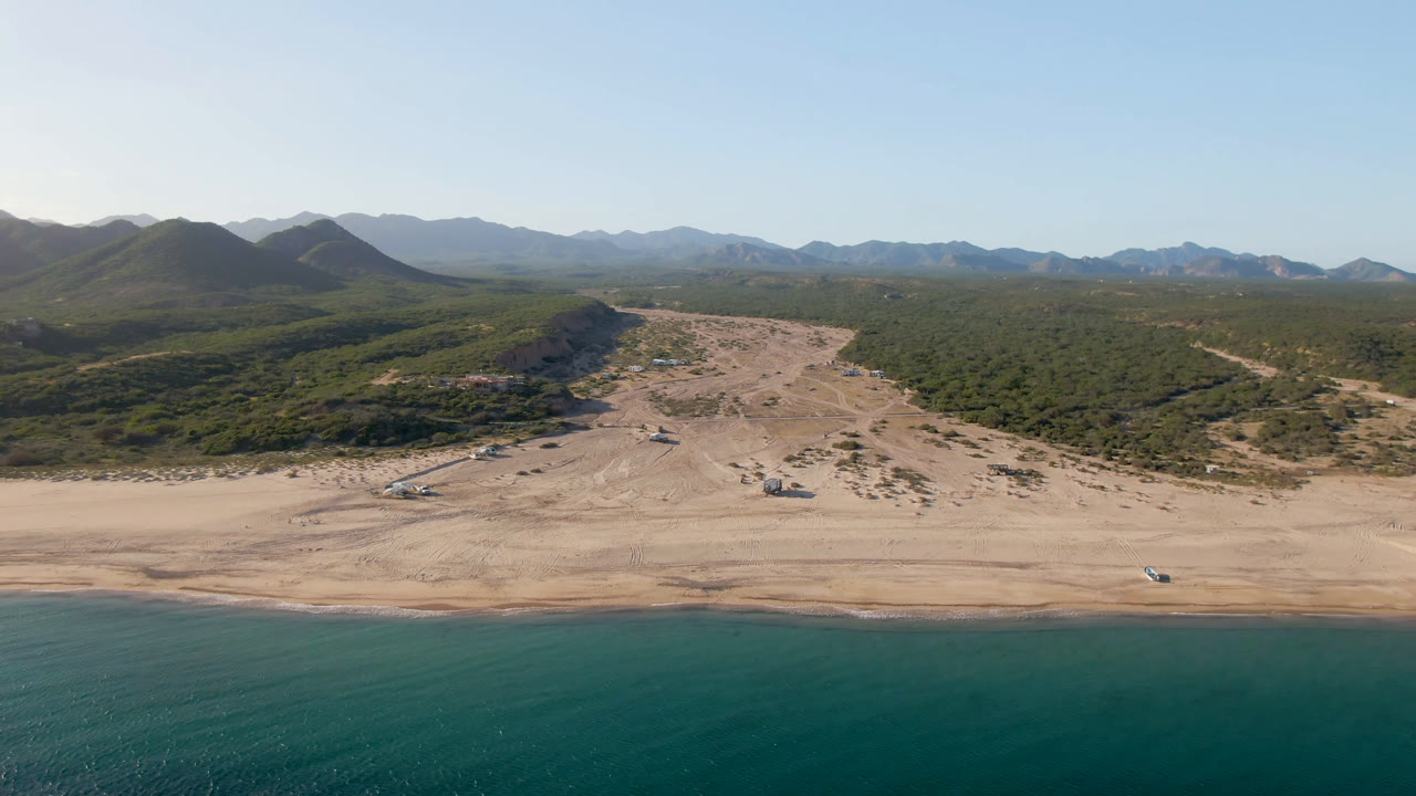 A high aerial view over Los Frailes shows teal water at the bottom edge, a broad sandy beach, scrub-covered hills, and distant mountains under clear daylight, with no visible people.
