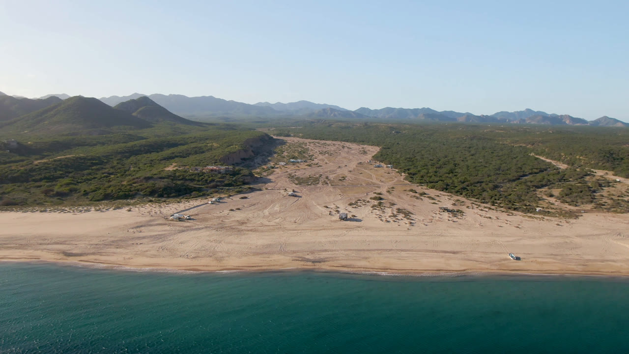 Los Frailes appears from above with the shoreline across the lower frame, pale sand and dune tracks through the center, green scrub on both sides, and no visible people.
