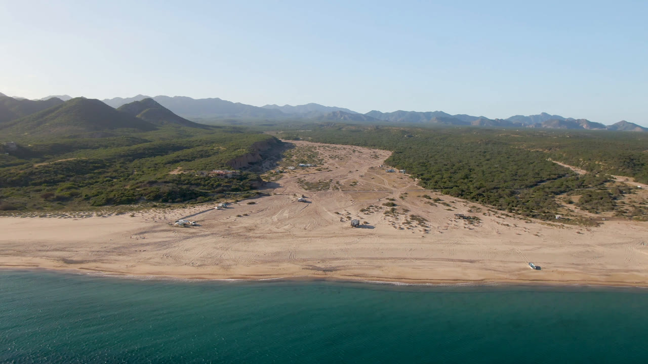 Aerial coastline with water, empty beach, low hills, and mountains.