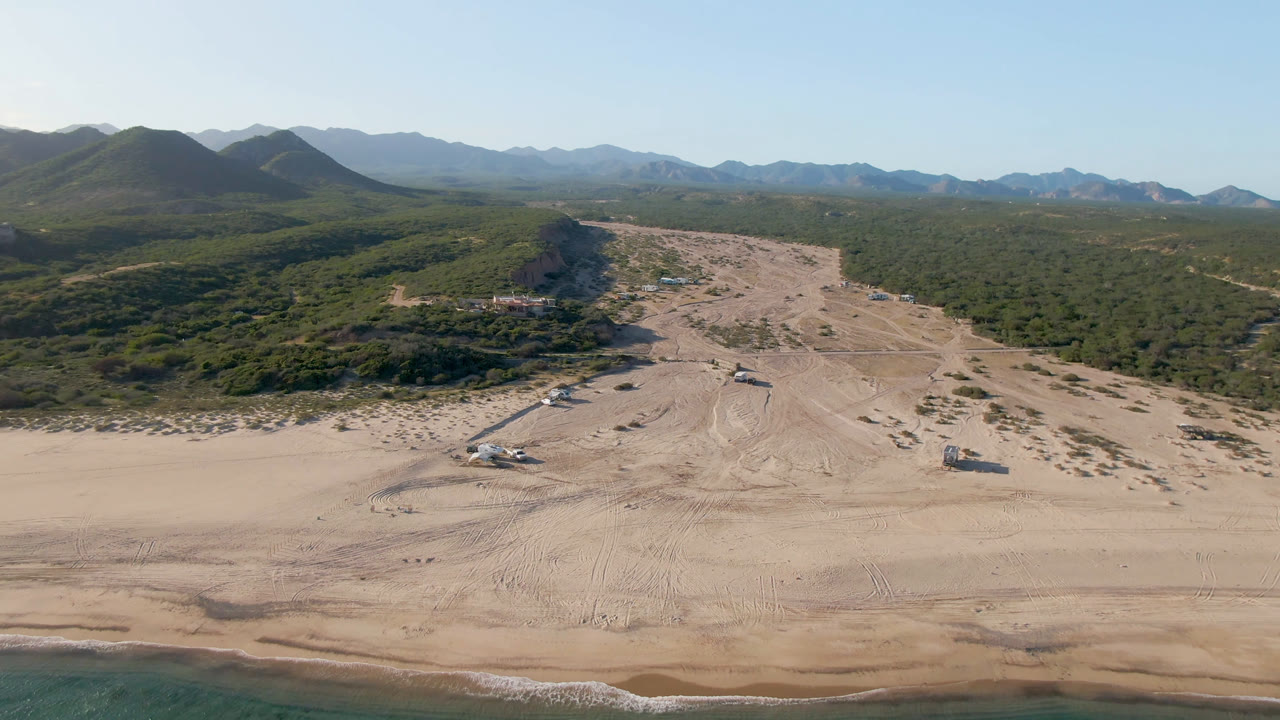 Wide aerial frame over Los Frailes, Baja California Sur, with sandy beach in front, green scrubland behind it, distant mountains, clear blue sky, and no visible people.