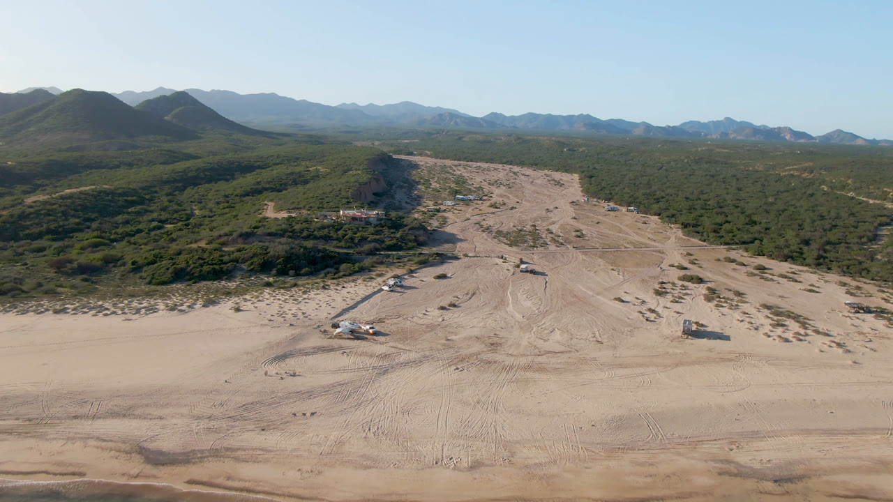 A second aerial view of Los Frailes shows more of the sand flats and track marks below, with scrub-covered terrain and a layered mountain horizon under clear sky, with no visible people.