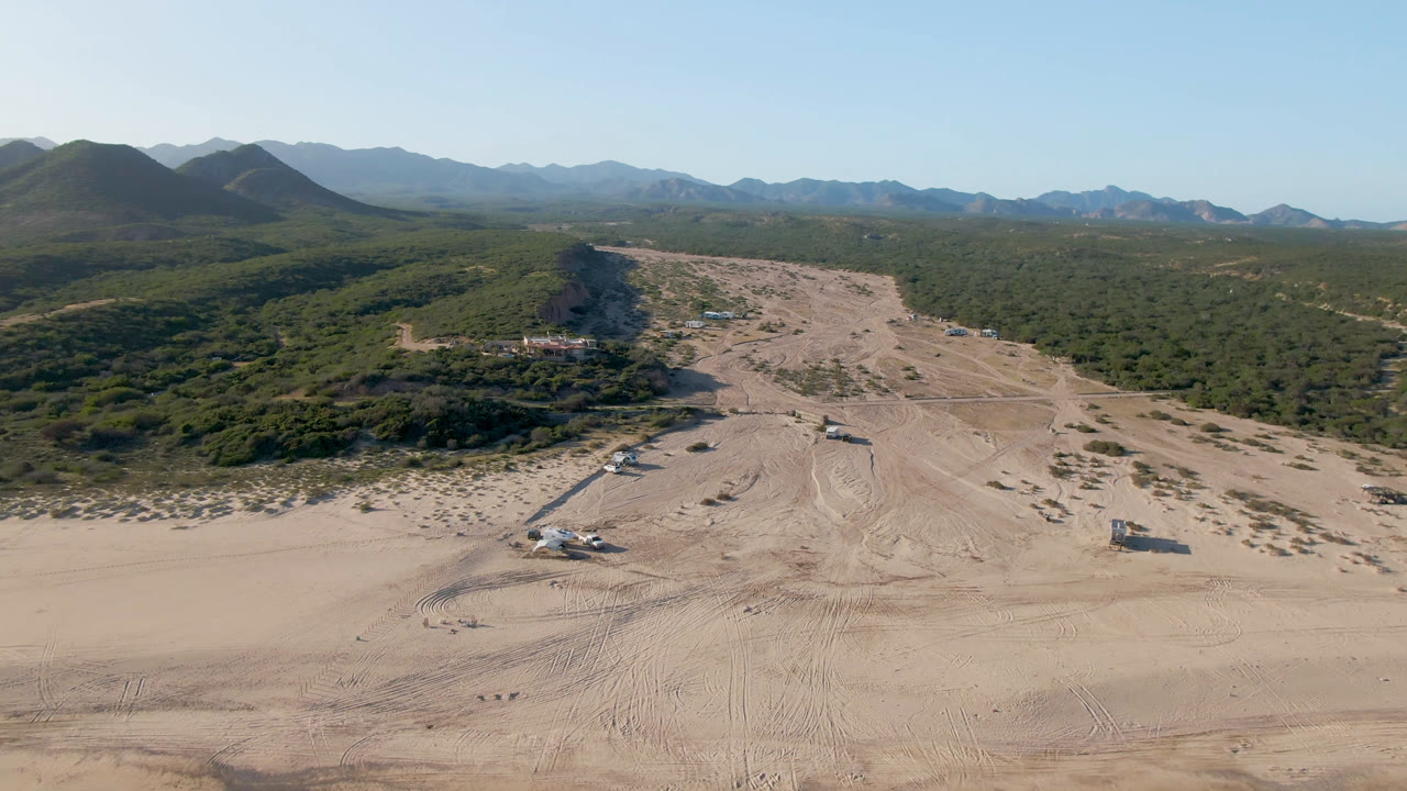 Drone view across a sandy shoreline toward scrubland and mountains.
