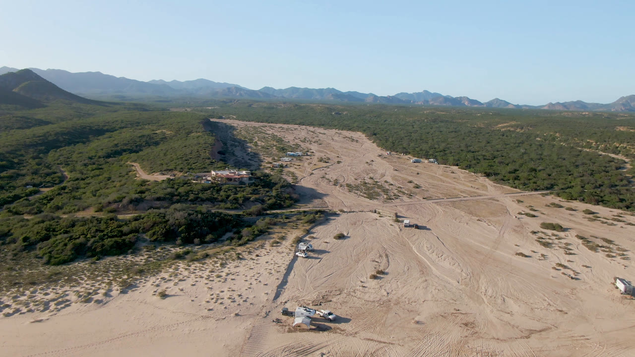 Aerial frame in Los Frailes, Baja California Sur, looking along a pale sandy wash with dirt tracks, clustered scrub, low buildings or vehicles, mountains, and no visible people.