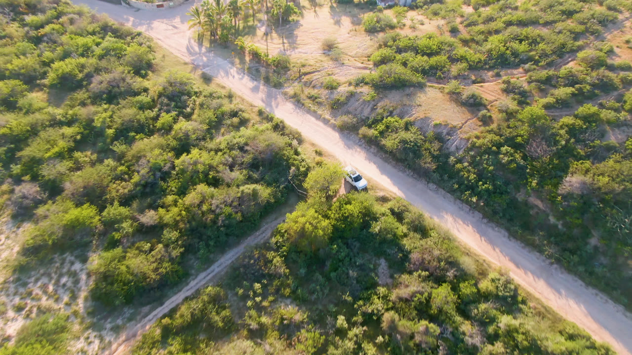 Aerial view in Los Frailes, Baja California Sur, Mexico, with a white car near the left side of a pale dirt road cutting through green scrub and sandy tracks, with no visible people.