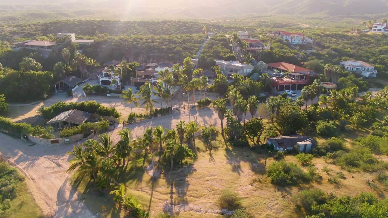 Aerial frame over Los Frailes, Baja California Sur, showing sandy roads, palm trees, scattered low homes, green vegetation, hazy hills, and no visible people.