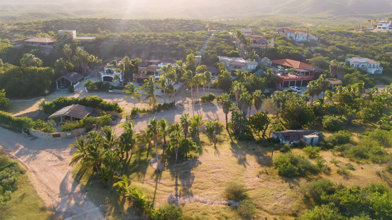 Drone-height view in Los Frailes with a broad sandy track in the foreground, palms casting long shadows, low buildings among vegetation, distant hills, and no visible people.