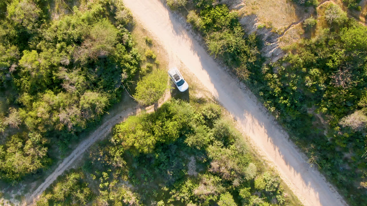 Overhead frame from Los Frailes showing the white vehicle slightly farther along the diagonal dirt road, bordered by thick green brush and no visible people.