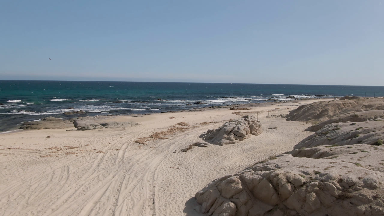 Wide view of Shipwrecks Beach in Los Cabos with pale sand, rounded rocks in the foreground, surf across the background, and no clearly visible people in this opening frame.