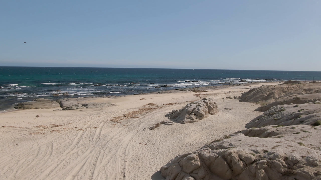Shipwrecks Beach shoreline with sand tracks leading toward low rock formations, blue-green ocean water, white surf, and a clear sky over the horizon.