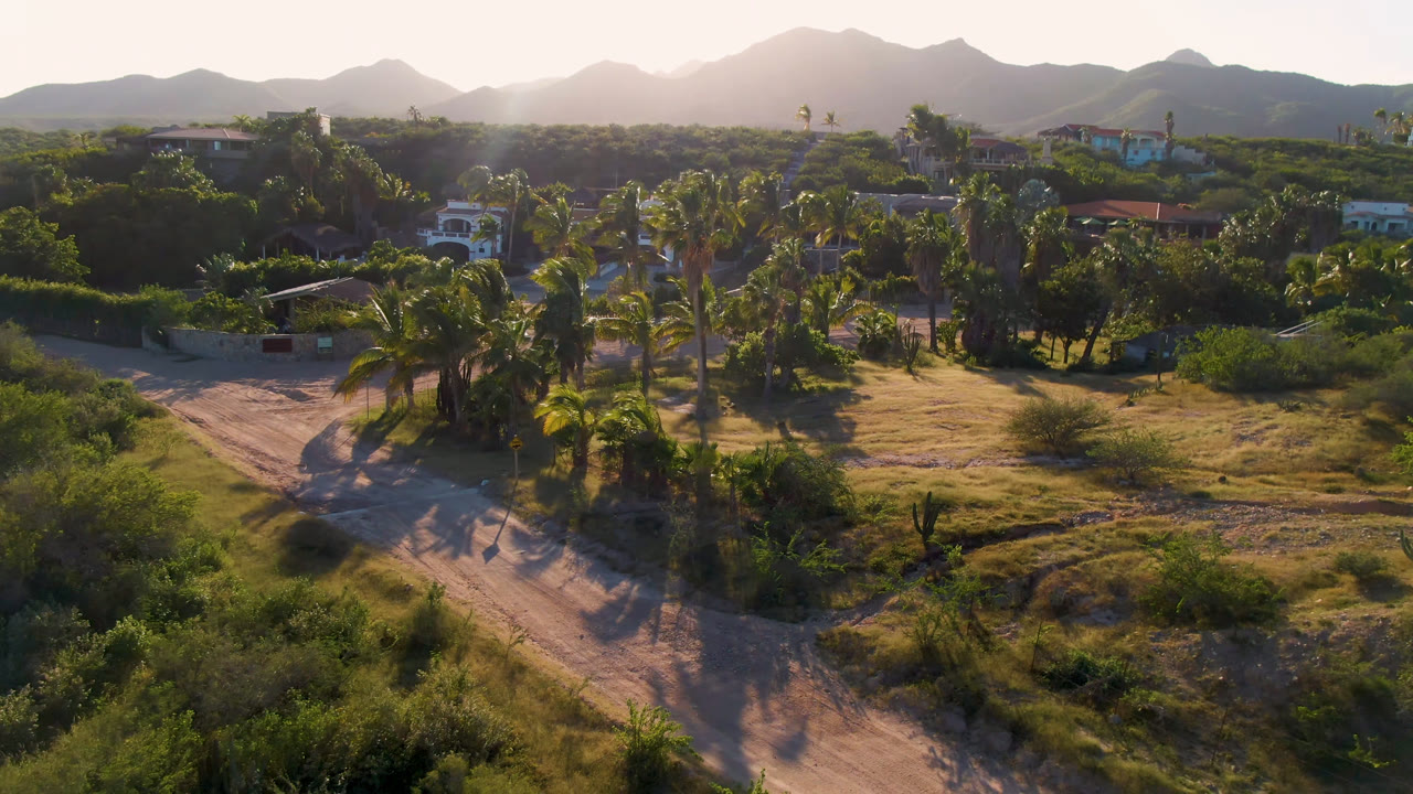 Los Frailes aerial view with the sandy road more centered below palm trees, low houses behind green vegetation, mountain ridges on the horizon, and no visible people.