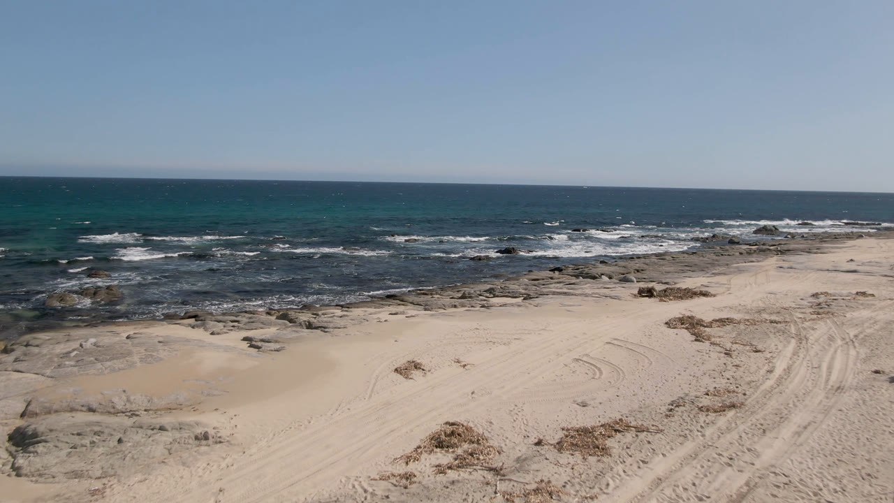 Wide view of Shipwrecks Beach in Los Cabos with pale sand, tire tracks, low rock shelves, white surf, blue ocean, clear sky, and no visible people.