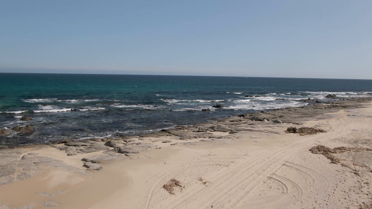 Shipwrecks Beach shoreline with darker shallow water around flat rocks, white wave foam across the midground, sand in the foreground, and no visible people.