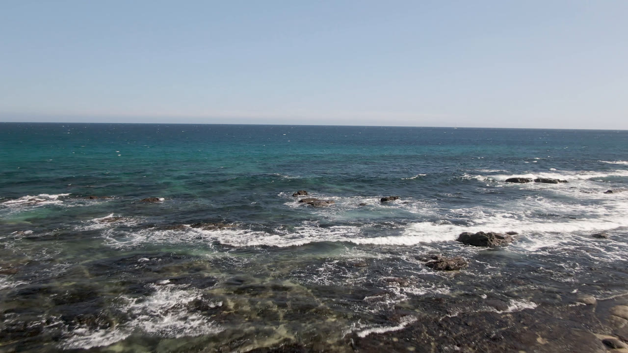 Elevated view over rocky water at Shipwrecks Beach in Los Cabos, with white wave foam in the foreground, turquoise patches offshore, a clear horizon, and no visible people.