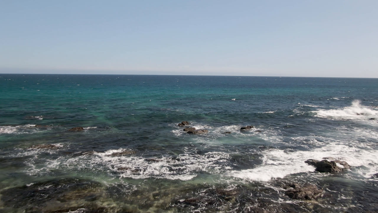 Shipwrecks Beach water fills the frame with scattered dark rocks, a fresh white wave break near the right side, green-blue shallows, and no visible people.