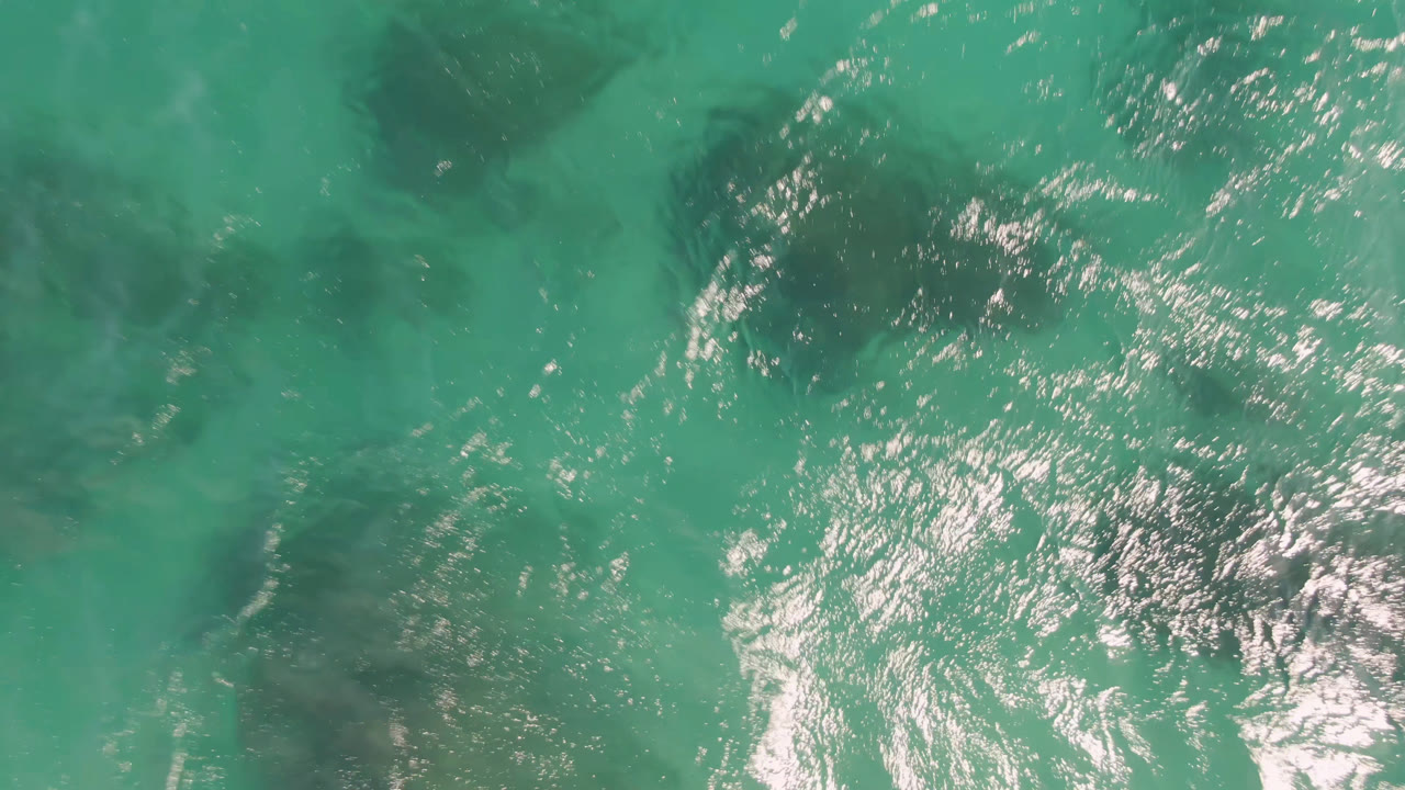 Overhead view of turquoise water at Shipwrecks Beach in Los Cabos, with bright sun glare on the left side and dark underwater patches visible, no visible people.