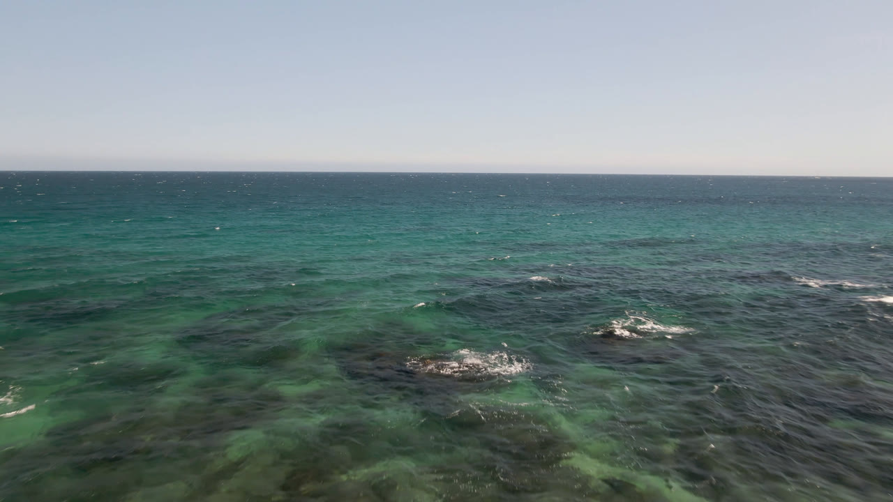 Wide view over the ocean at Shipwrecks Beach Los Cabos, with turquoise water, dark reef patches, small white breaks, a straight horizon, and no visible people.
