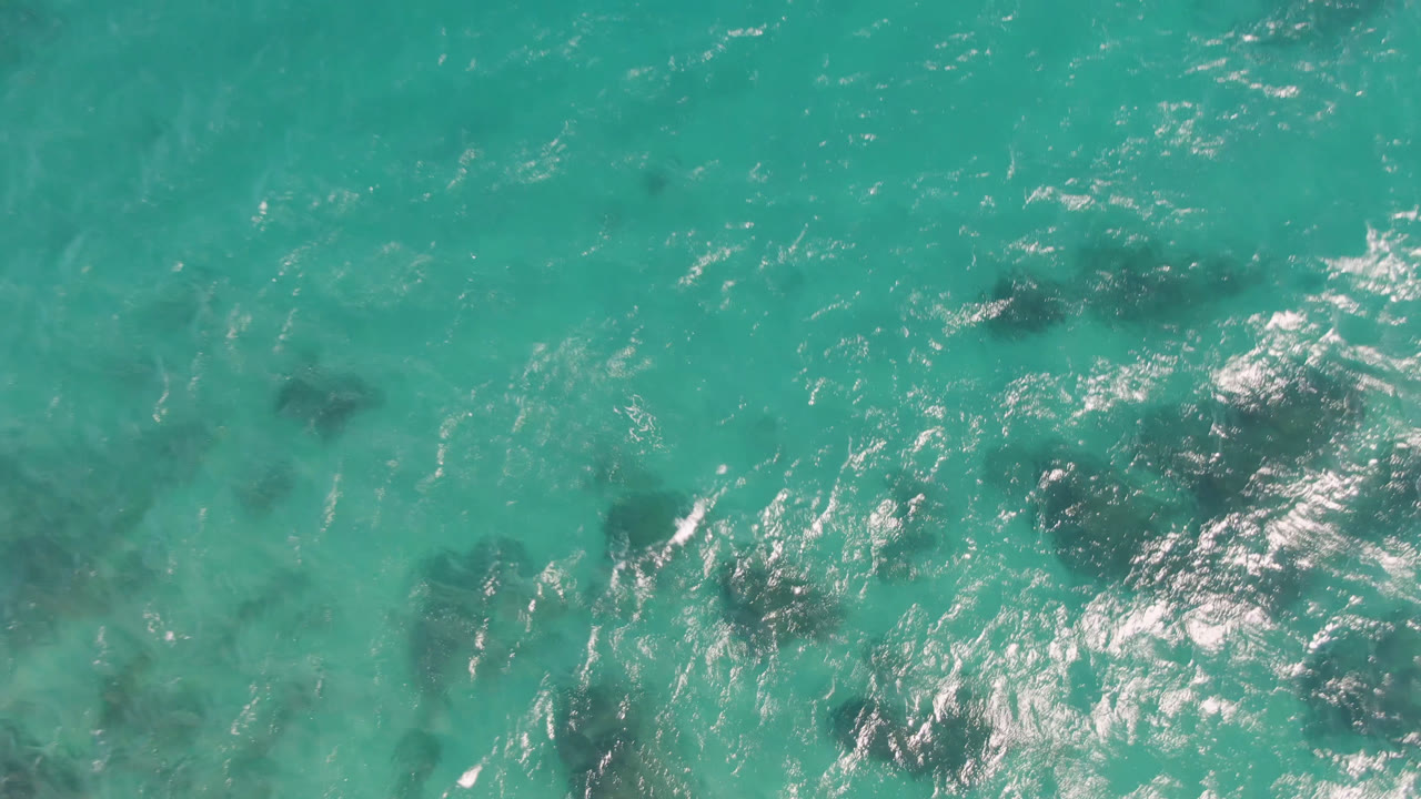Overhead view of turquoise water at Shipwrecks Beach in Los Cabos with dark submerged patches, light ripples, and no visible people.