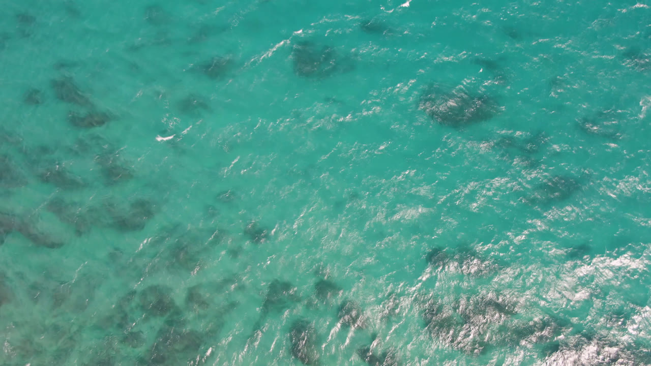 Aerial frame over Shipwrecks Beach water showing a denser cluster of dark submerged shapes near the center, fine ripples, and no visible people.