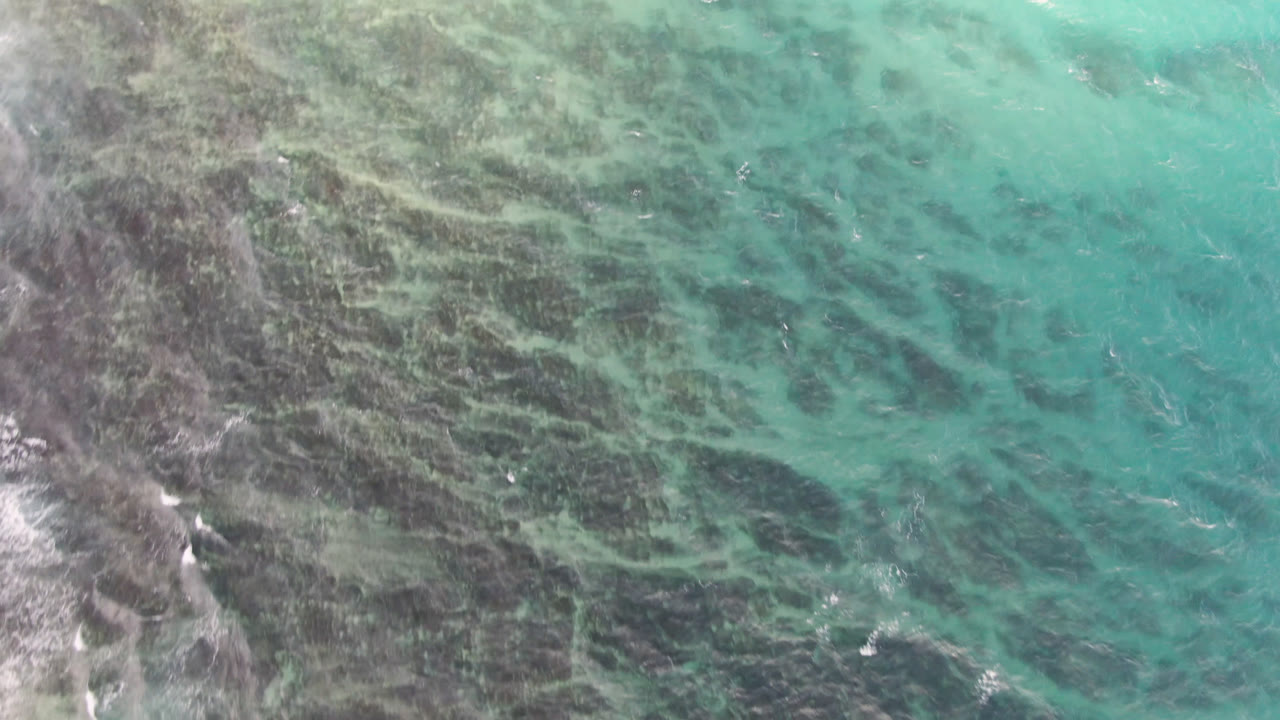 Aqua water spreads across dark submerged reef at Shipwrecks Beach in Los Cabos, with a few white wave flecks and no visible people.