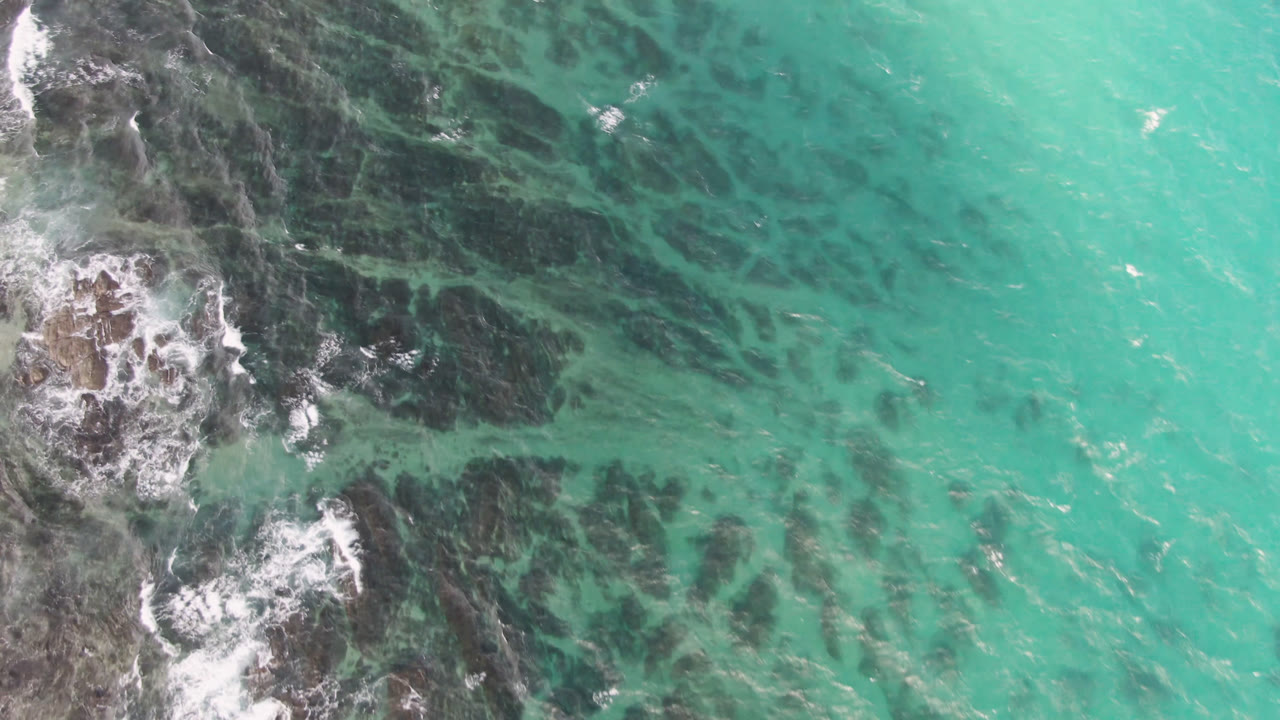 Opening overhead frame at Shipwrecks Beach Los Cabos shows turquoise-green water, dark submerged rock patterns, white foam on the left edge, and no visible people.