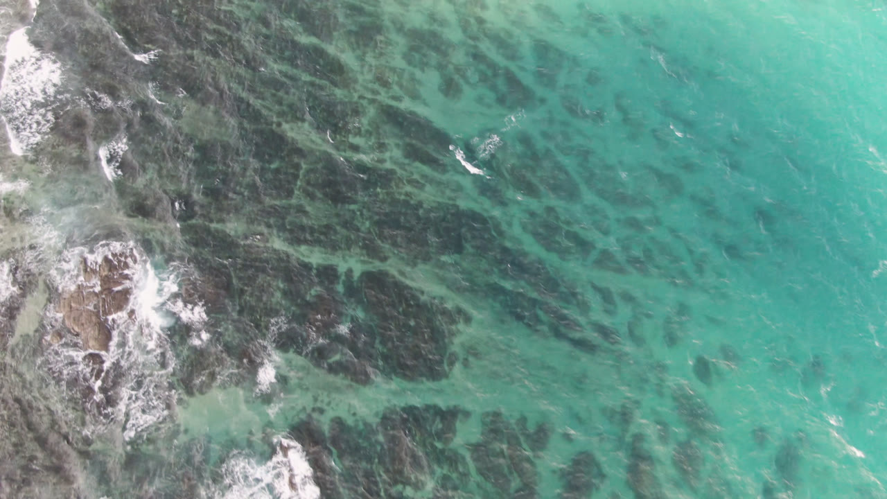 Overhead turquoise water with dark reef shapes and a foamy rock cluster at left.