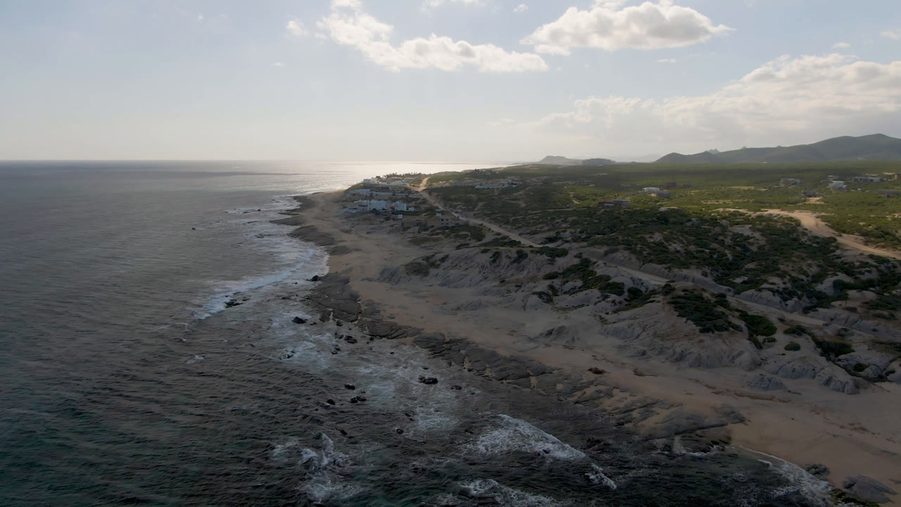 High aerial view over Shipwrecks Beach in Los Cabos, with dark ocean at left, pale sand and rocks below, green scrub inland, distant hills, bright clouds, and no visible people.