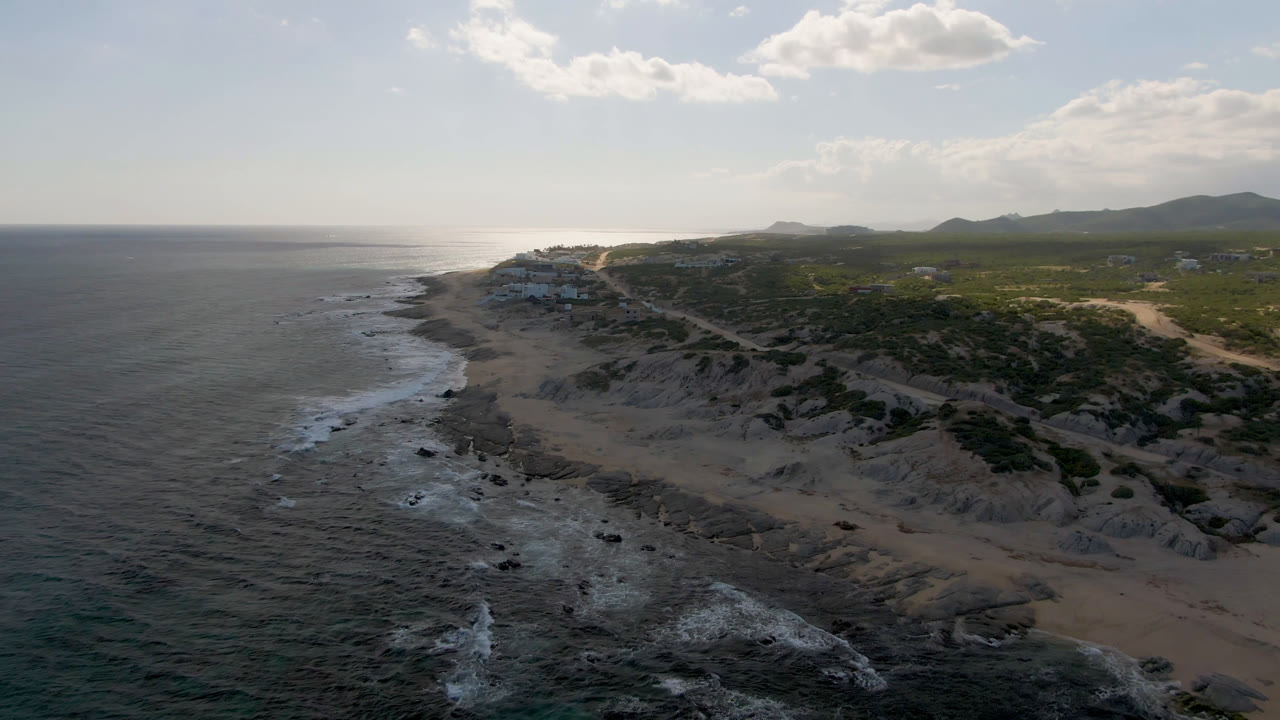 Aerial frame from Shipwrecks Beach in Los Cabos showing the ocean pushed farther across the left side, surf tracing the rocky shore, sandy cliff faces, inland scrub, and no visible people.