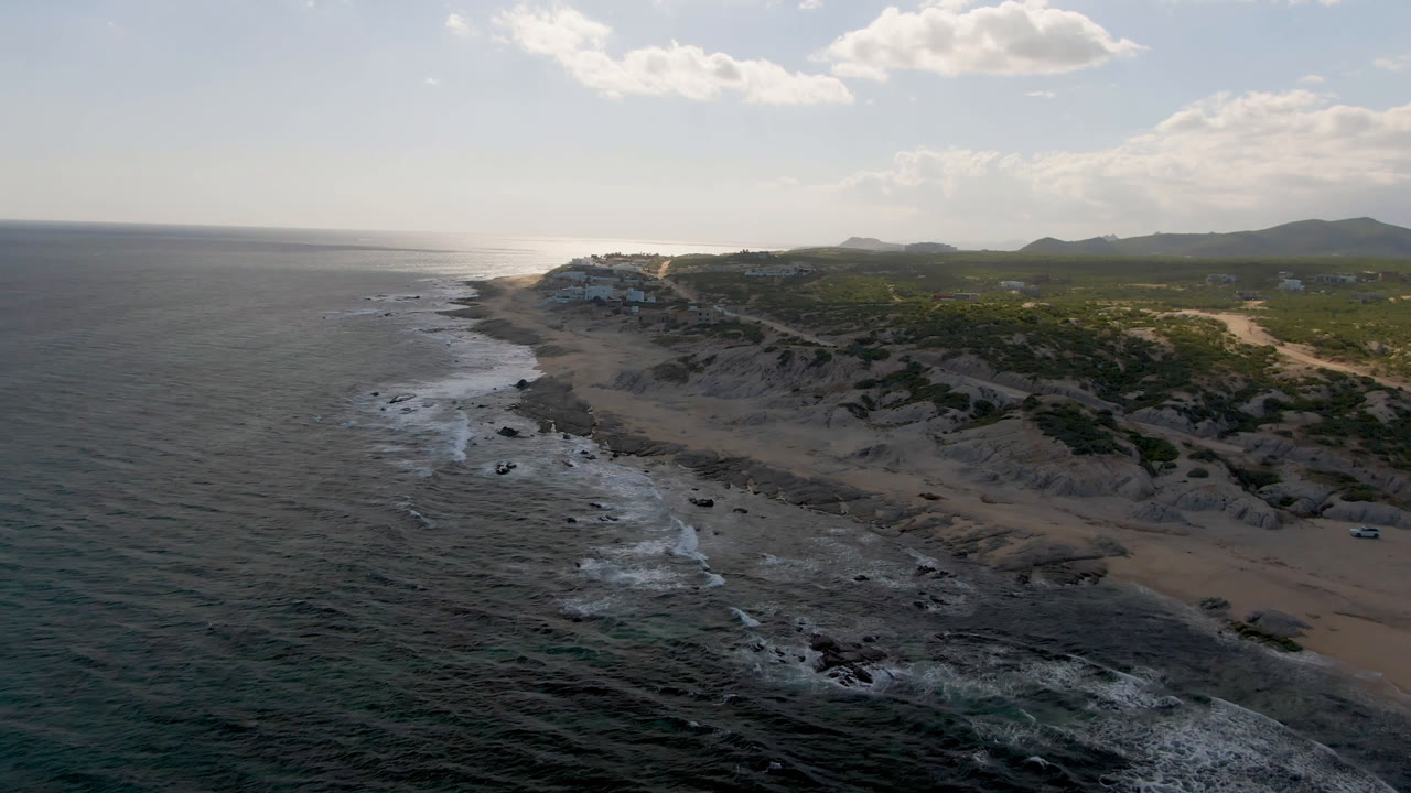Wide aerial view of Shipwrecks Beach in Los Cabos with ocean on the left, surf breaking over rocks, sandy dunes to the right, distant hills, bright clouds, and no visible people.