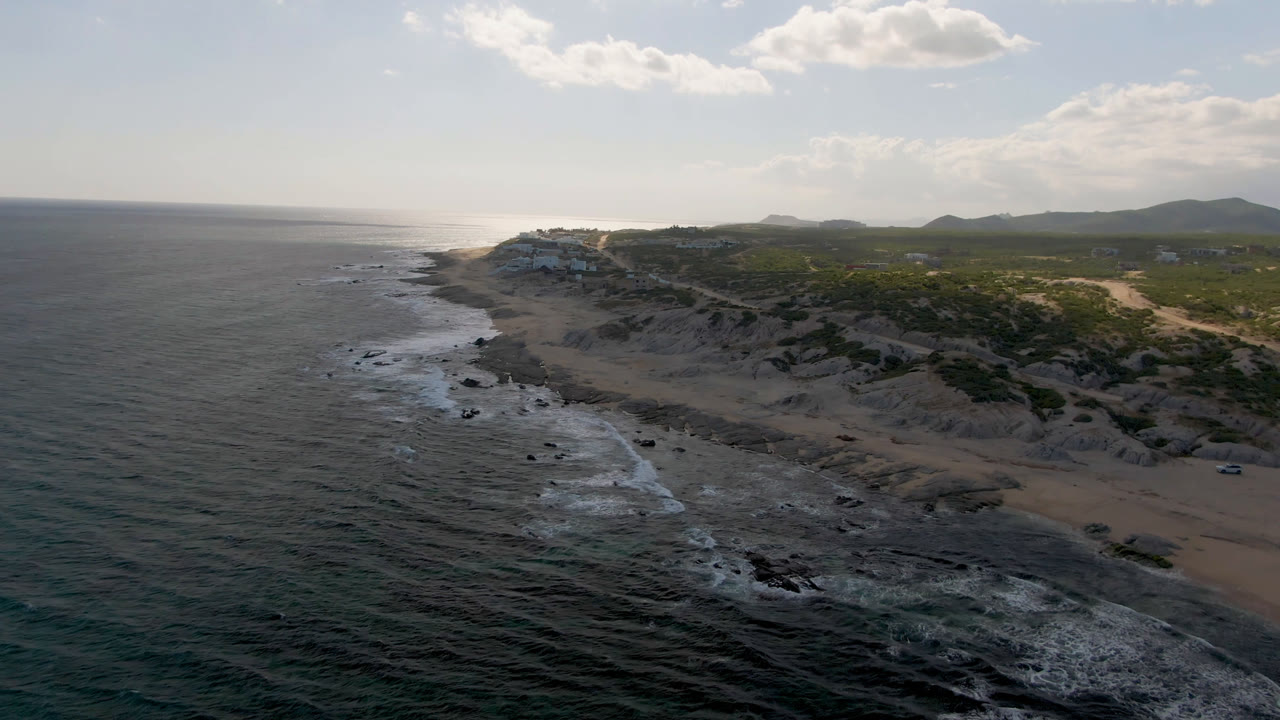 Aerial frame over Shipwrecks Beach Los Cabos showing a longer run of white water along the rocky shore, sand and dunes to the right, low hills beyond, and no visible people.