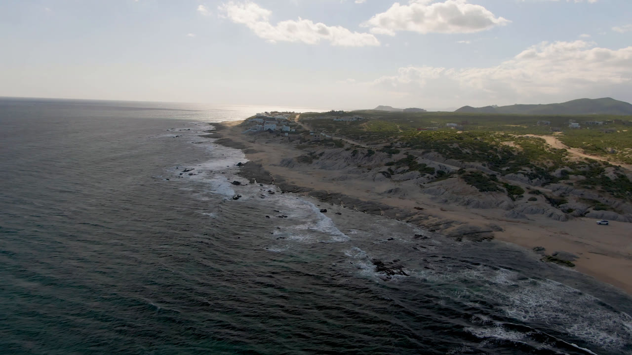 Aerial view of dark ocean water, foamy surf, beach dunes, and distant hills.