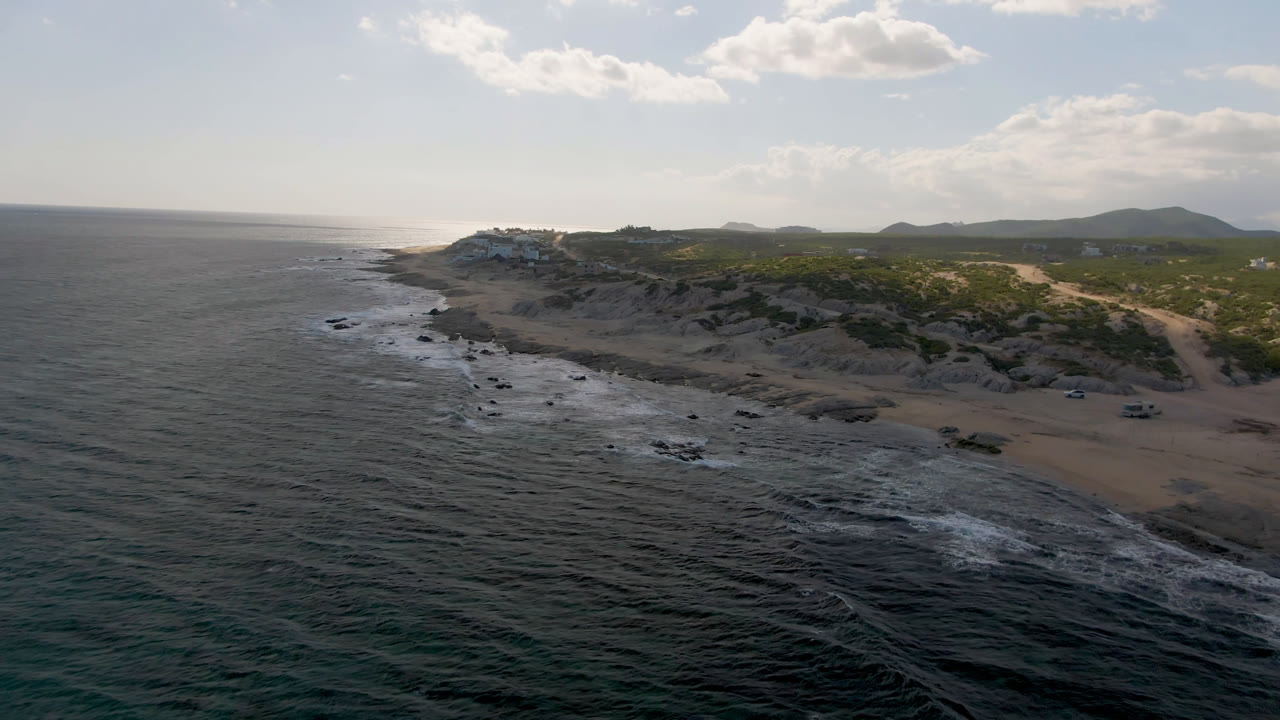 Aerial frame over Shipwrecks Beach in Los Cabos, with dark ocean in the foreground, surf along rocks and sand, scrubland behind the beach, distant hills, and no visible people.