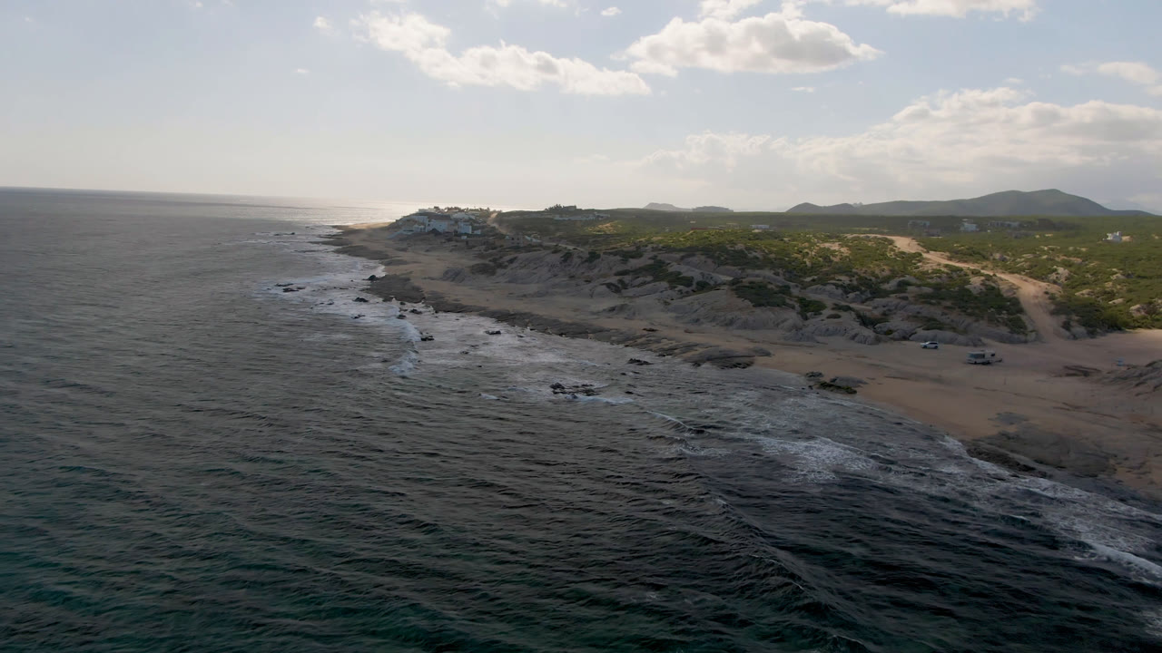 Wide aerial view at Shipwrecks Beach in Los Cabos, showing ocean on the left, a strip of white surf, sandy beach and rocks in the middle, green scrub and hills in the distance, with no visible people.