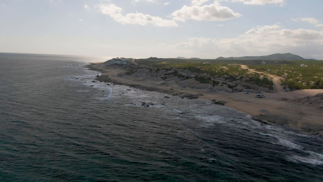 Aerial coastline view with white waves over dark water and a sandy beach beyond.
