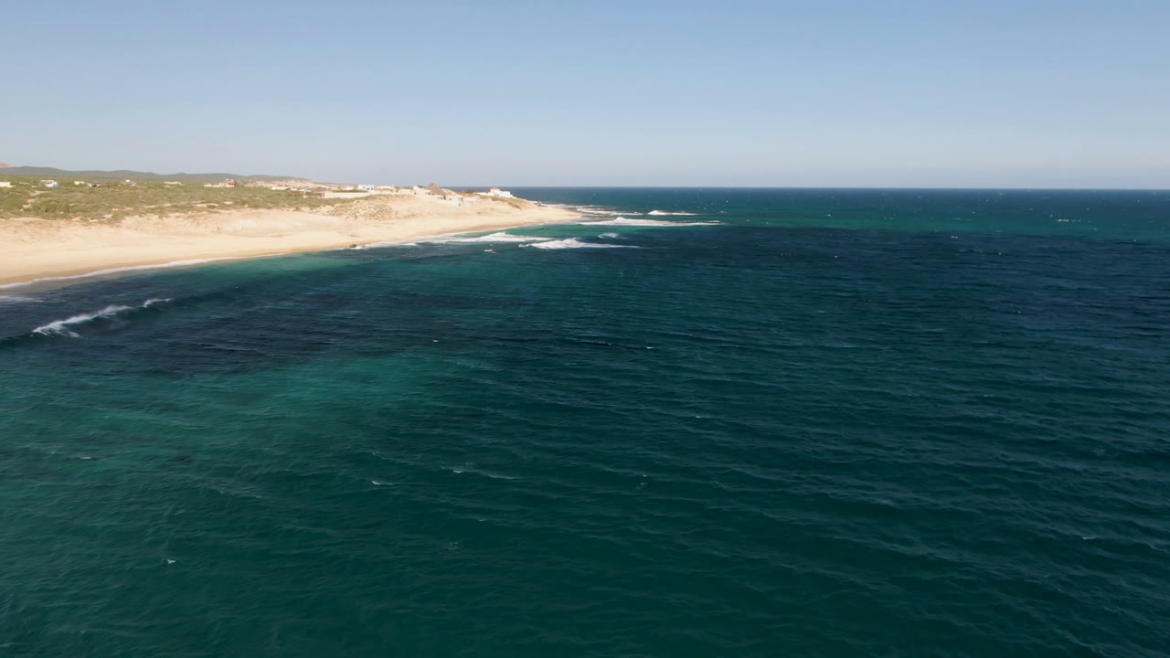Opening aerial frame over Shipwrecks Beach Los Cabos, with dark teal water in the foreground, pale sand across the shore, low green dunes beyond, and no visible people.