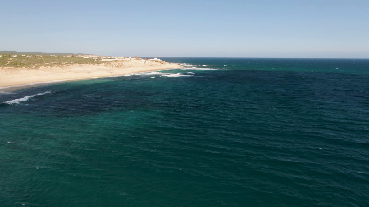 Aerial frame at Shipwrecks Beach Los Cabos showing rippled dark water, a curving white surf line, pale sand, low coastal vegetation, and no visible people.