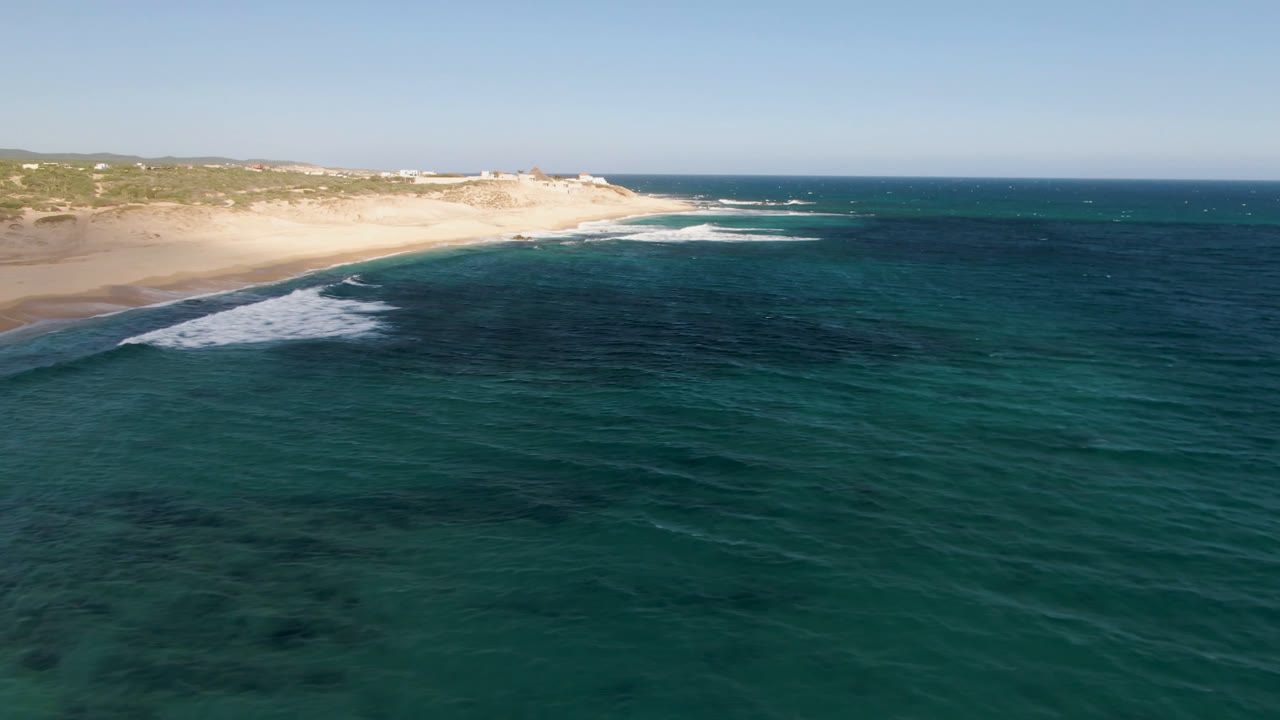 Opening frame of Shipwrecks Beach in Los Cabos with teal water in the foreground, a white surf line along the sand, dunes beyond the shore, and no visible people.