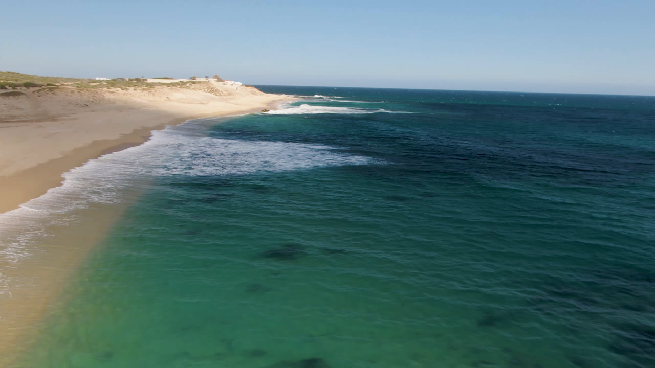 Aerial view of Shipwrecks Beach Los Cabos with turquoise water in the foreground, a pale sandy shoreline curving left, low dunes beyond, and no visible people.
