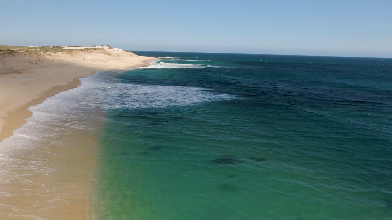 Shipwrecks Beach Los Cabos seen from above with the beach curving through the center, pale foam along the shore, darker blue water offshore, and no visible people.