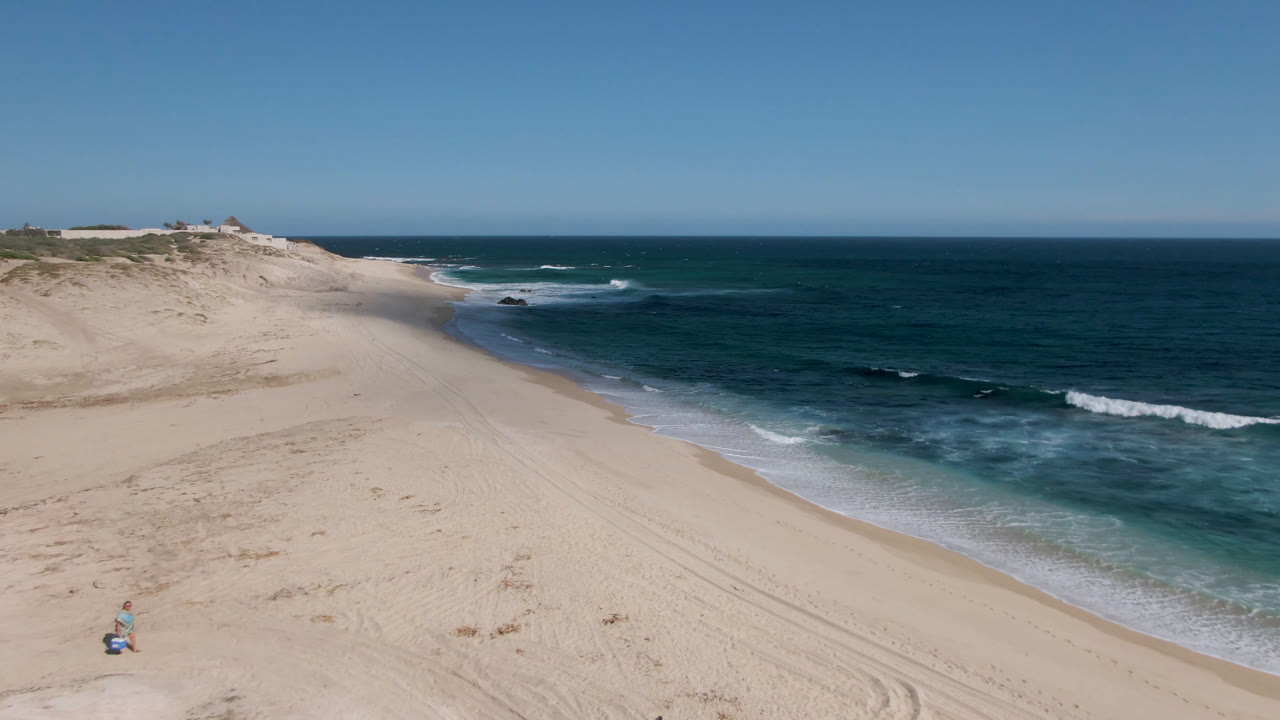 Aerial view of sandy beach, blue ocean, surf, and clear sky.