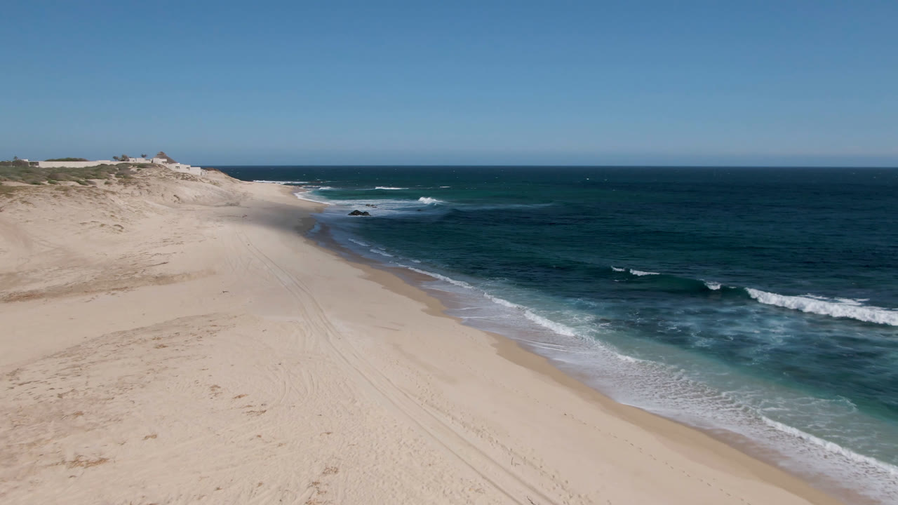 Drone view along Shipwrecks Beach in Los Cabos with wide empty sand, a curving wet shoreline, small white breakers, deep blue water, and no visible people.